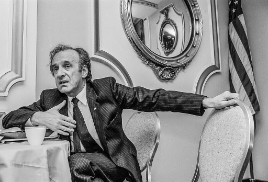 Man seated at a table, speaking and gesturing with his hands. He's wearing a suit, and an American flag is in the background.